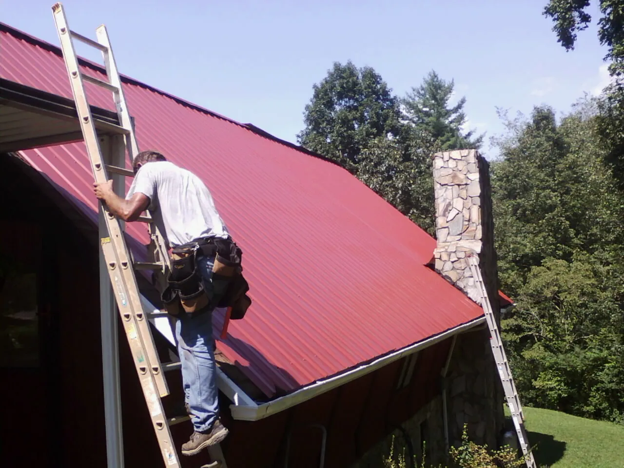 Roofer on a ladder beside a red metal roof and stone chimney — Soffit & Fascia Repair by Roofing Brothers in Fredericktown
